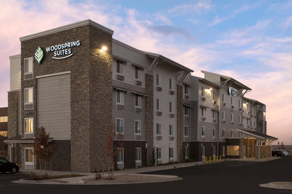 A four-story WoodSpring Suites hotel building with stone and gray siding exterior, illuminated signage, and a small parking area, pictured at dusk with a colorful sky in the background.