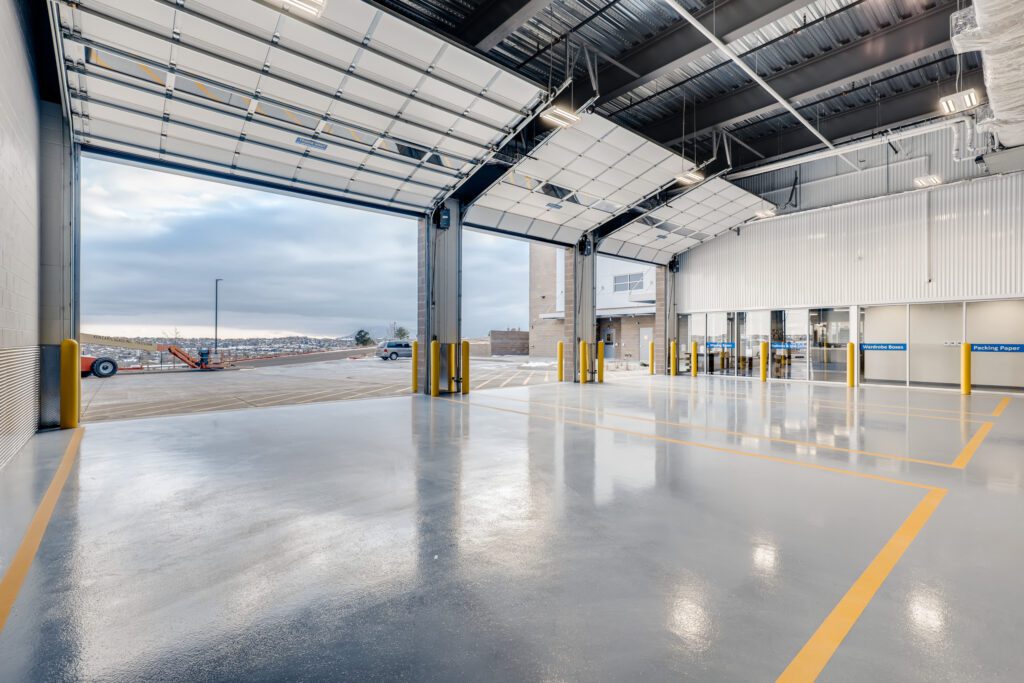Spacious, clean industrial garage with shiny gray floors, high ceilings, and large open bay doors. Yellow lines mark lanes on the floor, and glass doors lead to an interior area. Outdoor view shows a cloudy sky and some snow.