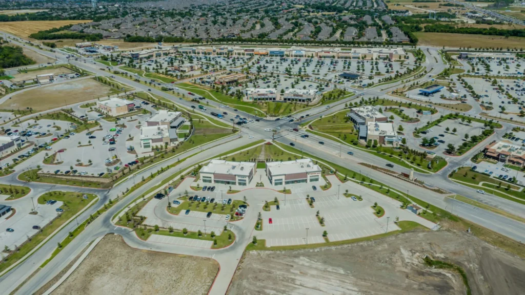 Aerial view of a suburban commercial area with large parking lots, intersecting roads, multiple shopping centers, and residential neighborhoods in the background. Some parking lots and areas are partially empty.