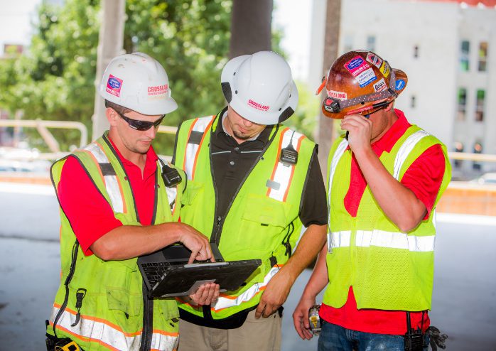 Three construction workers wearing PPE as they stand outside and look at laptop.