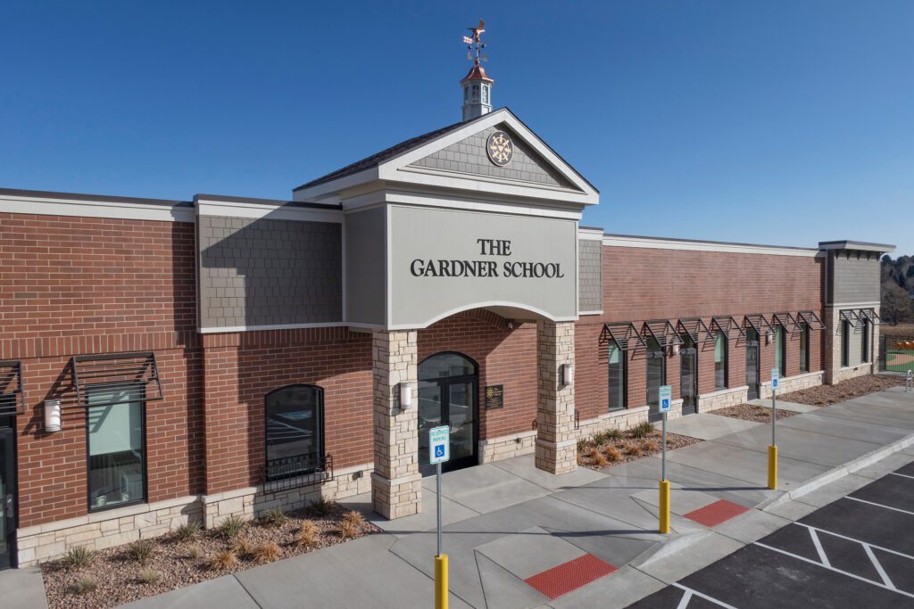 A modern brick building with a sign reading "The Gardner School" above the entrance, featuring large windows, a clock tower, and an accessible parking area in front under a clear blue sky.