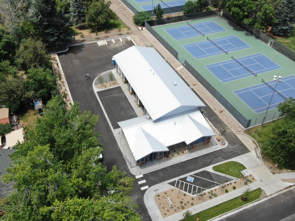 Aerial view of a building with a white roof next to multiple blue and green tennis courts, surrounded by trees, parking areas, and paved walkways.