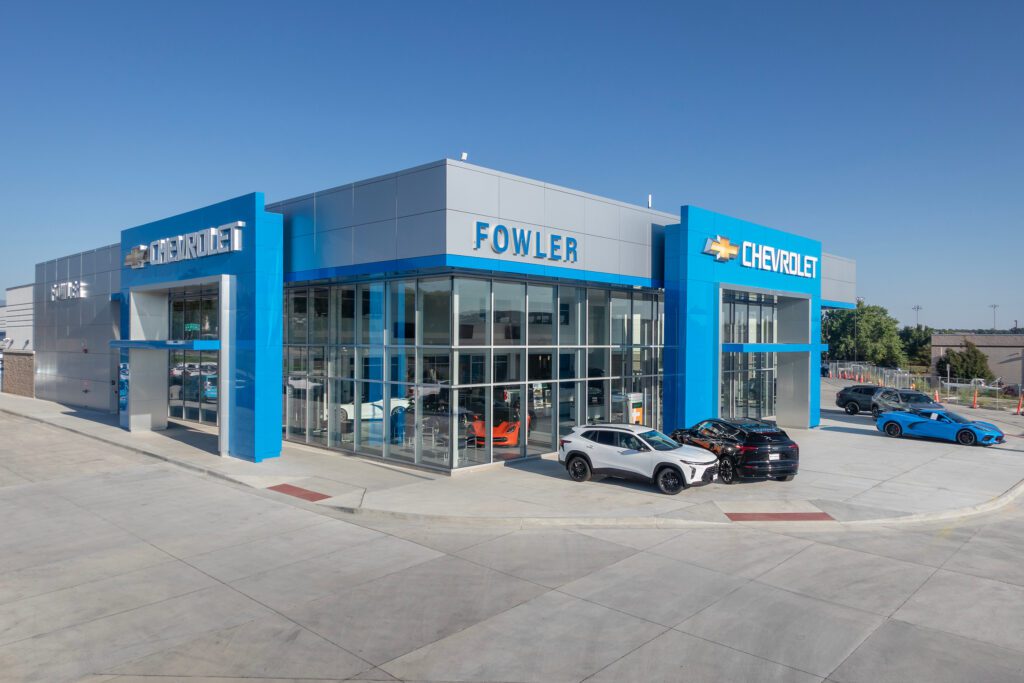 Modern Chevrolet dealership building with large glass windows, blue accents, and "Fowler" and "Chevrolet" signs. Several cars, including black and white models, are parked in front under a clear blue sky.