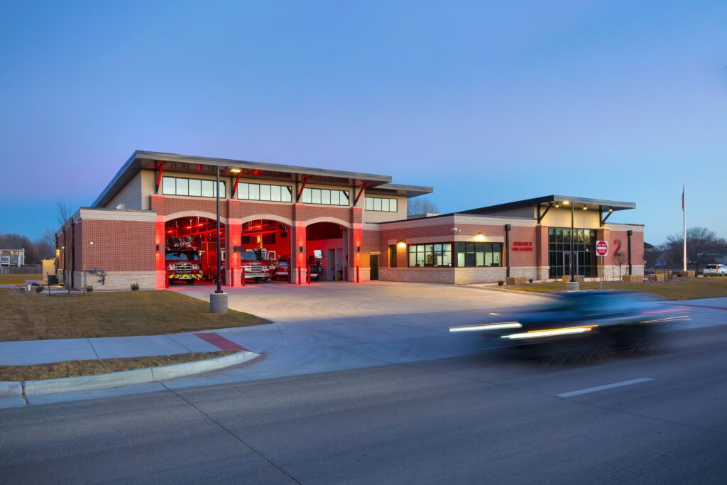 A modern brick fire station with three open garage bays, each illuminated with red lights, sits near a road at dusk. A fire truck is visible inside, and a blurred vehicle passes by in the foreground.