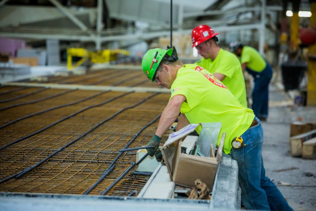 Construction workers in high-visibility Crossland Prefab shirts and hard hats assemble a reinforced steel framework for a prefabricated concrete panel. One worker in a green hard hat carefully adjusts the placement of rebar and conduit, while others in the background contribute to the production process in an industrial facility.