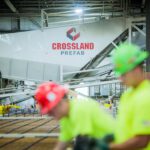 Two construction workers in high-visibility shirts and hard hats work in the foreground of a Crossland Prefab facility. The background features industrial equipment and a large Crossland Prefab sign, emphasizing the prefab construction process.