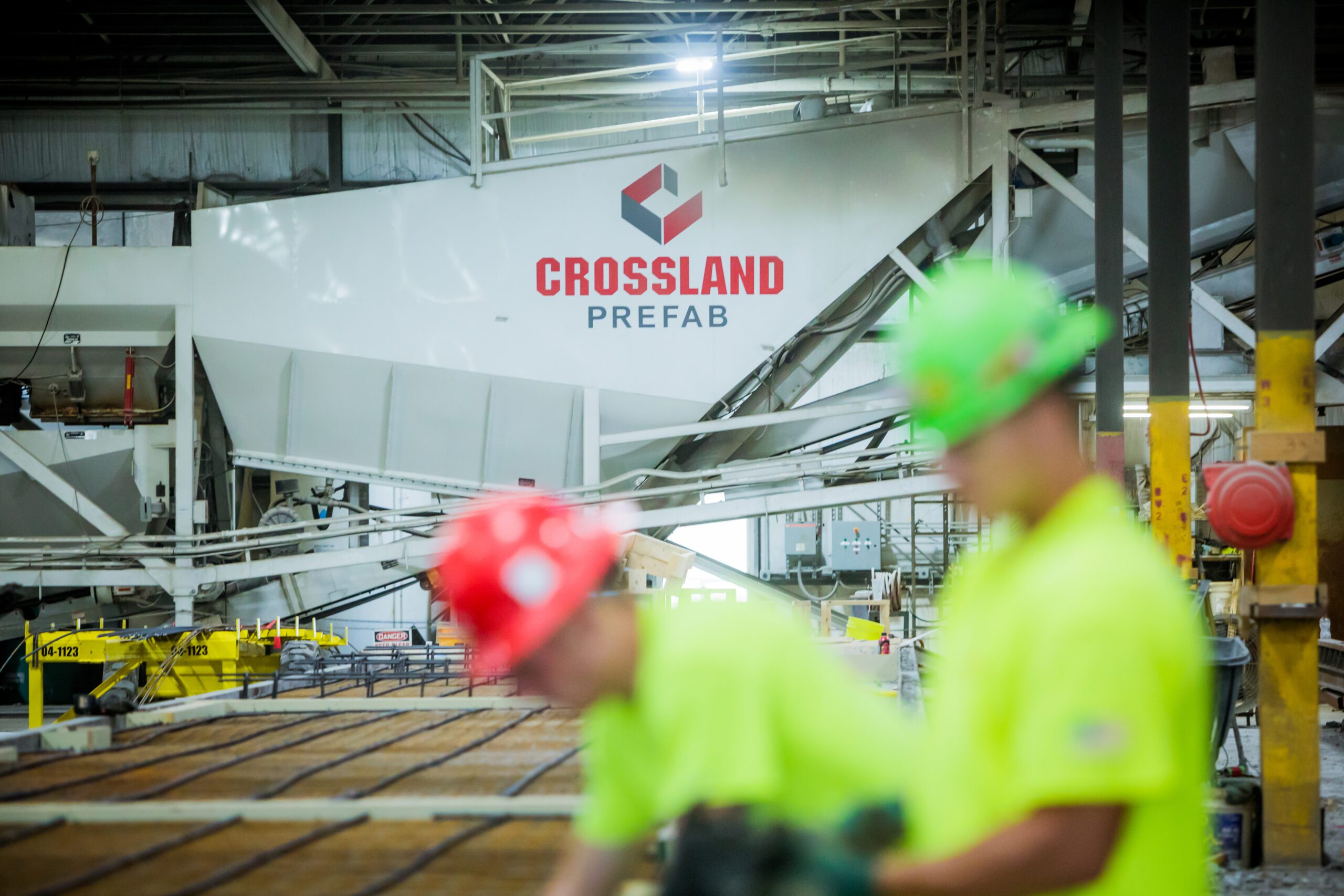 Two construction workers in high-visibility shirts and hard hats work in the foreground of a Crossland Prefab facility. The background features industrial equipment and a large Crossland Prefab sign, emphasizing the prefab construction process.