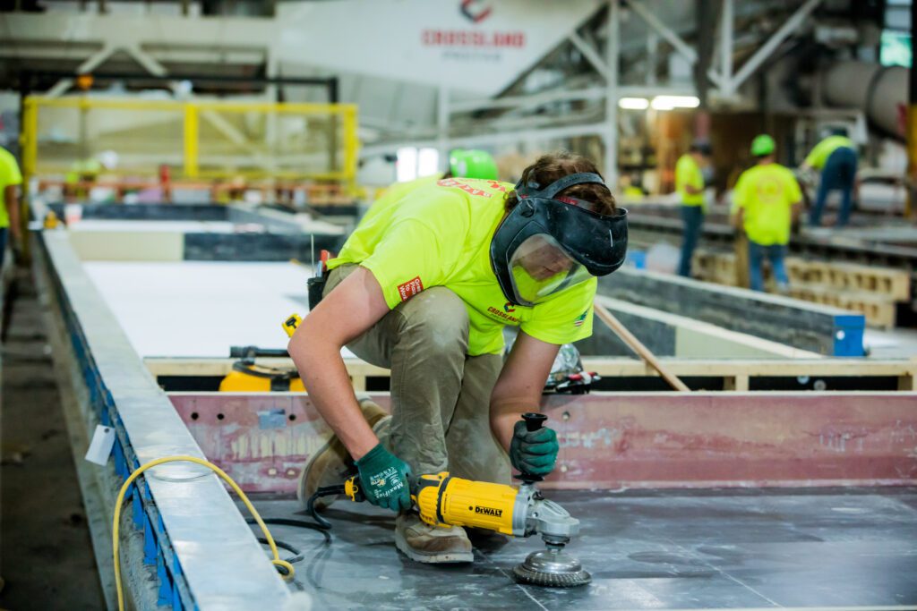 A worker in a high-visibility Crossland Prefab shirt and protective face shield uses a DeWalt power tool to grind or polish a surface at a prefab construction facility. Other team members in safety gear are working in the background, highlighting the active production environment.