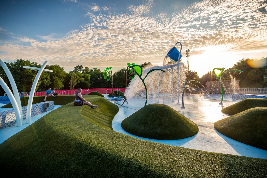 Children play at a splash pad with water features, spraying and pouring water, surrounded by green artificial turf mounds. The sun sets in the background, casting a warm glow and creating a partly cloudy sky.
