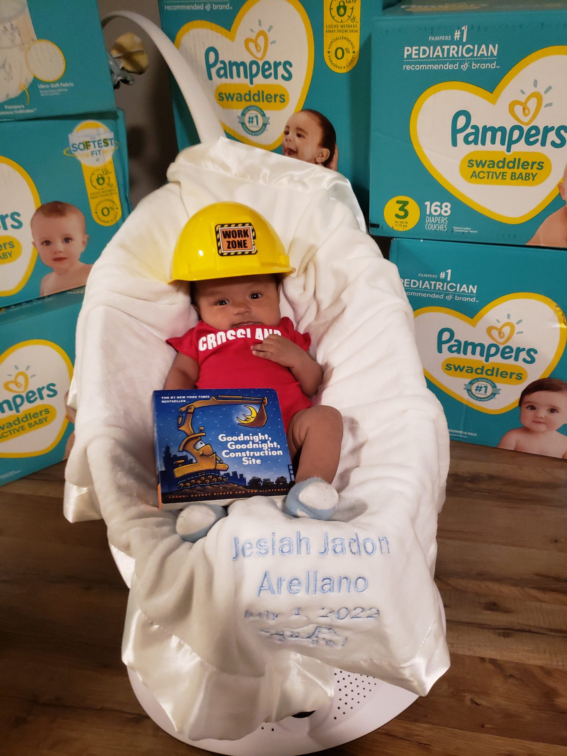 A baby wearing a hard hat in front of boxes of pampers.