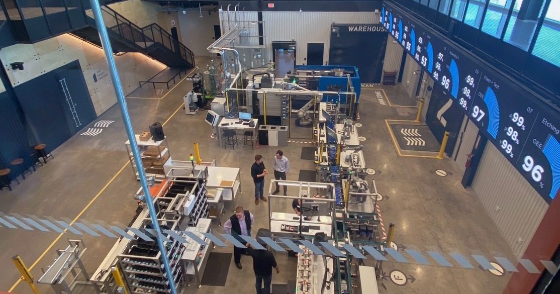 A few people stand and talk in a spacious, modern factory or lab with various industrial machines, workstations, and large digital displays showing data on the wall.