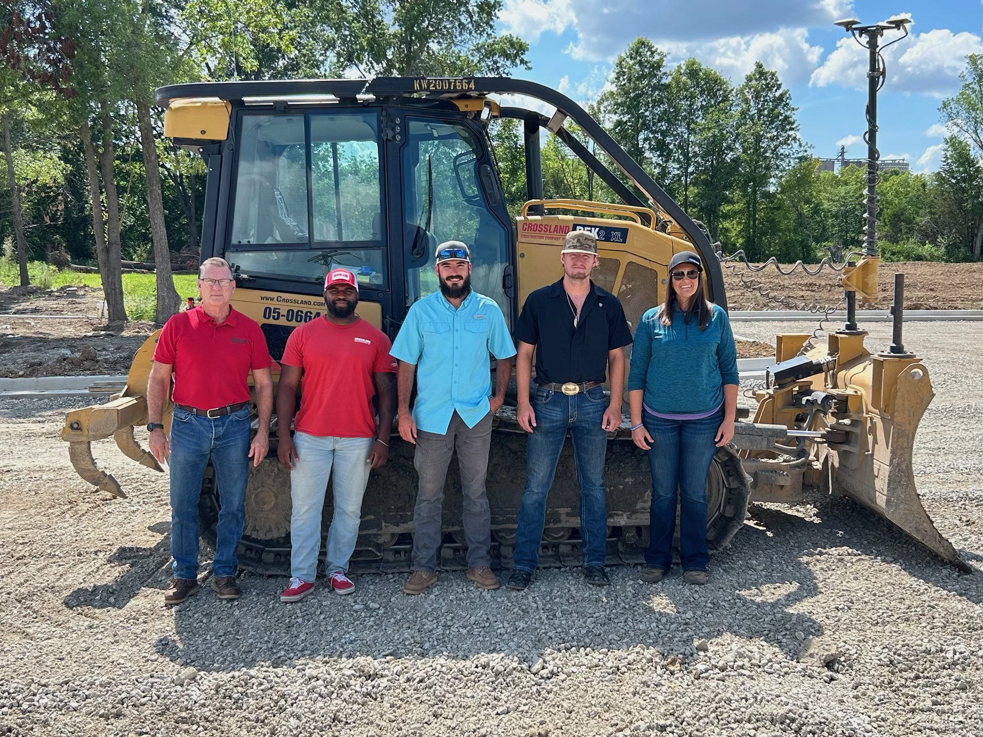 Five people stand in front of a large bulldozer on a construction site, with trees and a blue sky in the background. Three men wear hats, and everyone is dressed in casual attire.