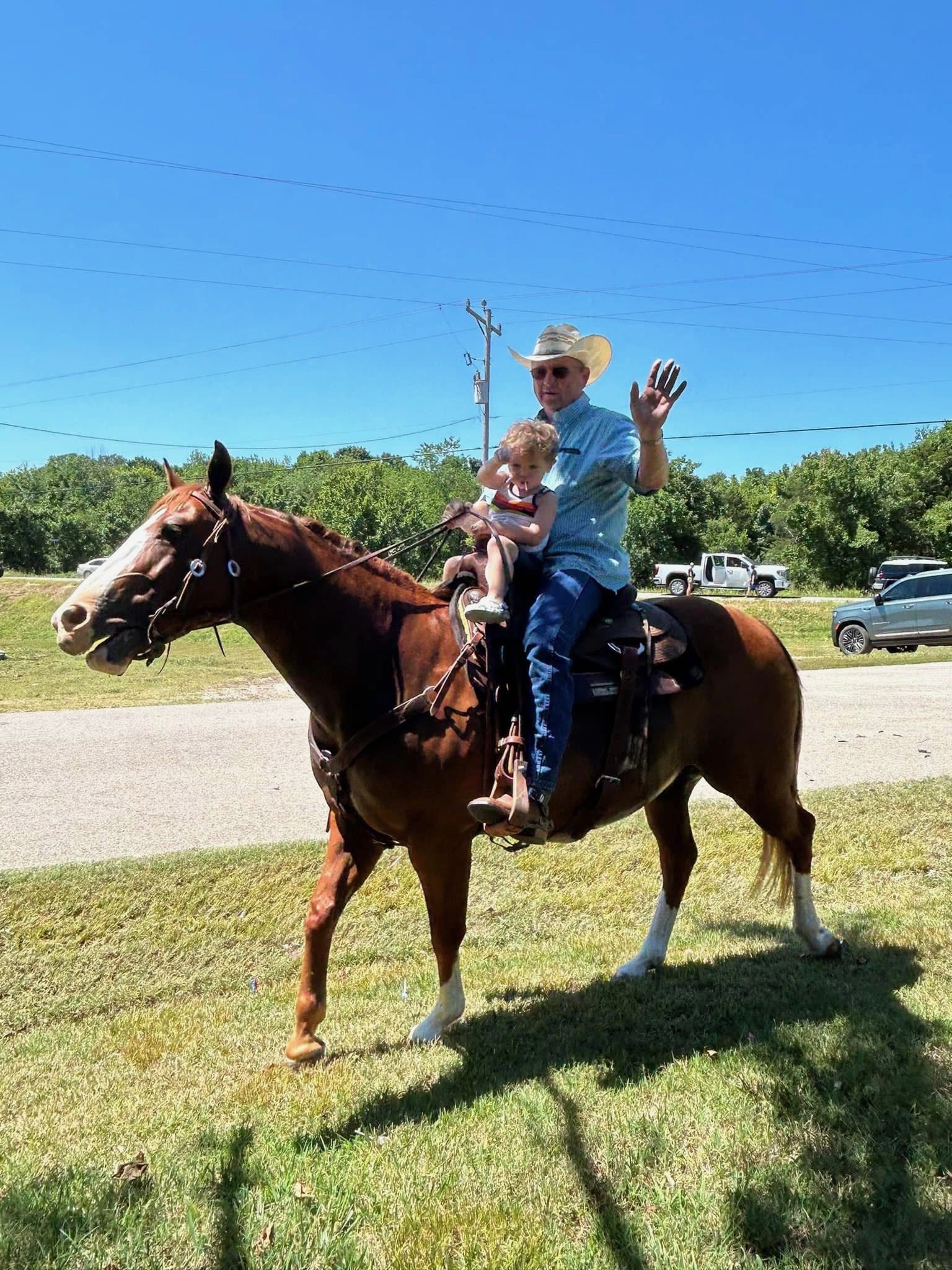 An older man in a cowboy hat waves while riding a brown horse with a small child sitting in front of him. They are outdoors on a grassy area with a road, cars, and trees in the background under a clear blue sky.