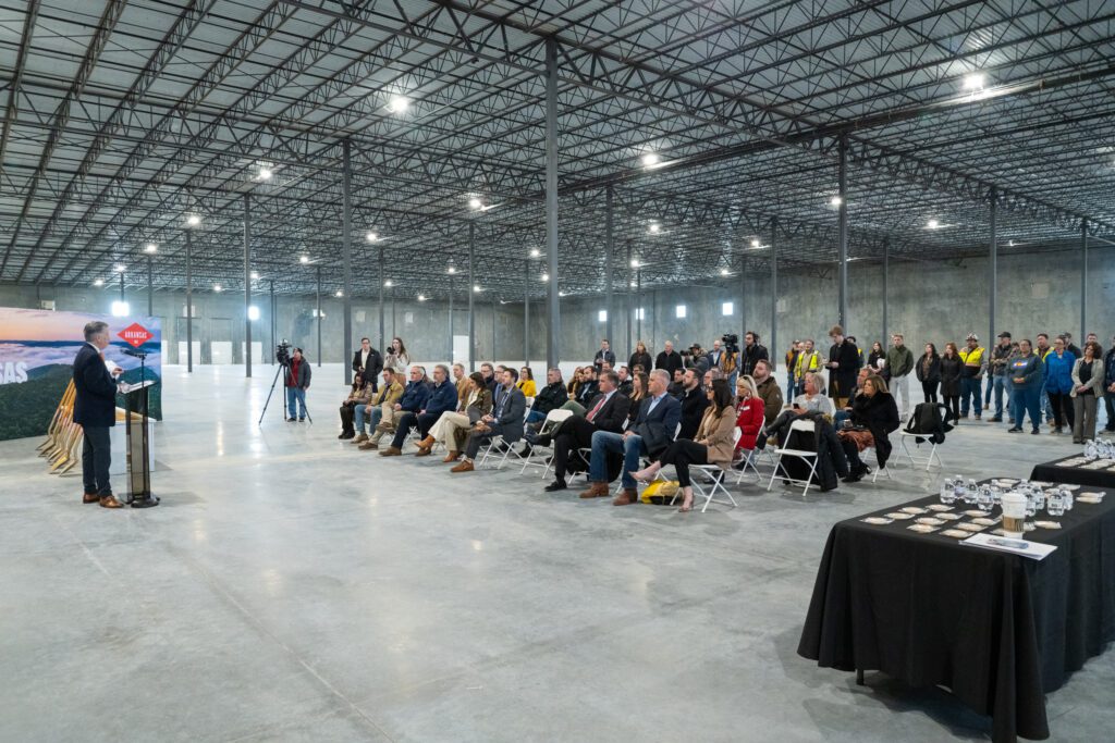 A large gathering inside a spacious industrial building listens to a speaker at a podium during a groundbreaking or dedication ceremony. Attendees, including business professionals, construction workers, and community members, are seated in rows of chairs while others stand in the background. A banner featuring an Arkansas landscape is positioned near the speaker, and a table with branded materials is visible in the foreground. The unfinished warehouse-like space has exposed steel framework and a polished concrete floor, highlighting the scale of the project.







