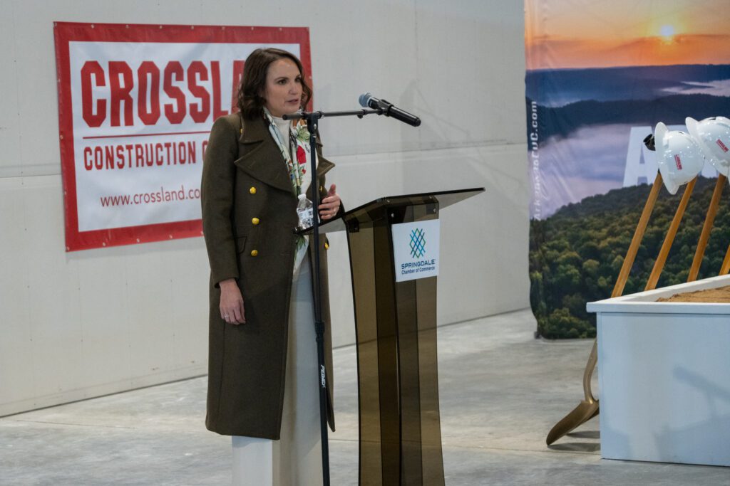 A woman stands at a podium delivering a speech during a groundbreaking event. The podium bears the logo of the Springdale Chamber of Commerce, and behind her, a Crossland Construction banner is displayed. To the right, white hard hats rest on shovels, positioned in a sandbox for the ceremonial groundbreaking. A backdrop featuring a scenic landscape adds to the event’s atmosphere.