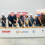 A group of ten people wearing hard hats and holding shovels participate in an indoor groundbreaking ceremony. They are standing behind a rectangular sand-filled box with the logos of Crossland Construction, Keytronic, Integrated Consulting Engineers, Inc., and Adams Architecture displayed on the front. A Crossland Construction banner is visible in the background, along with posters and a scenic backdrop. The participants are smiling and tossing sand with their shovels, celebrating the start of a new construction project.