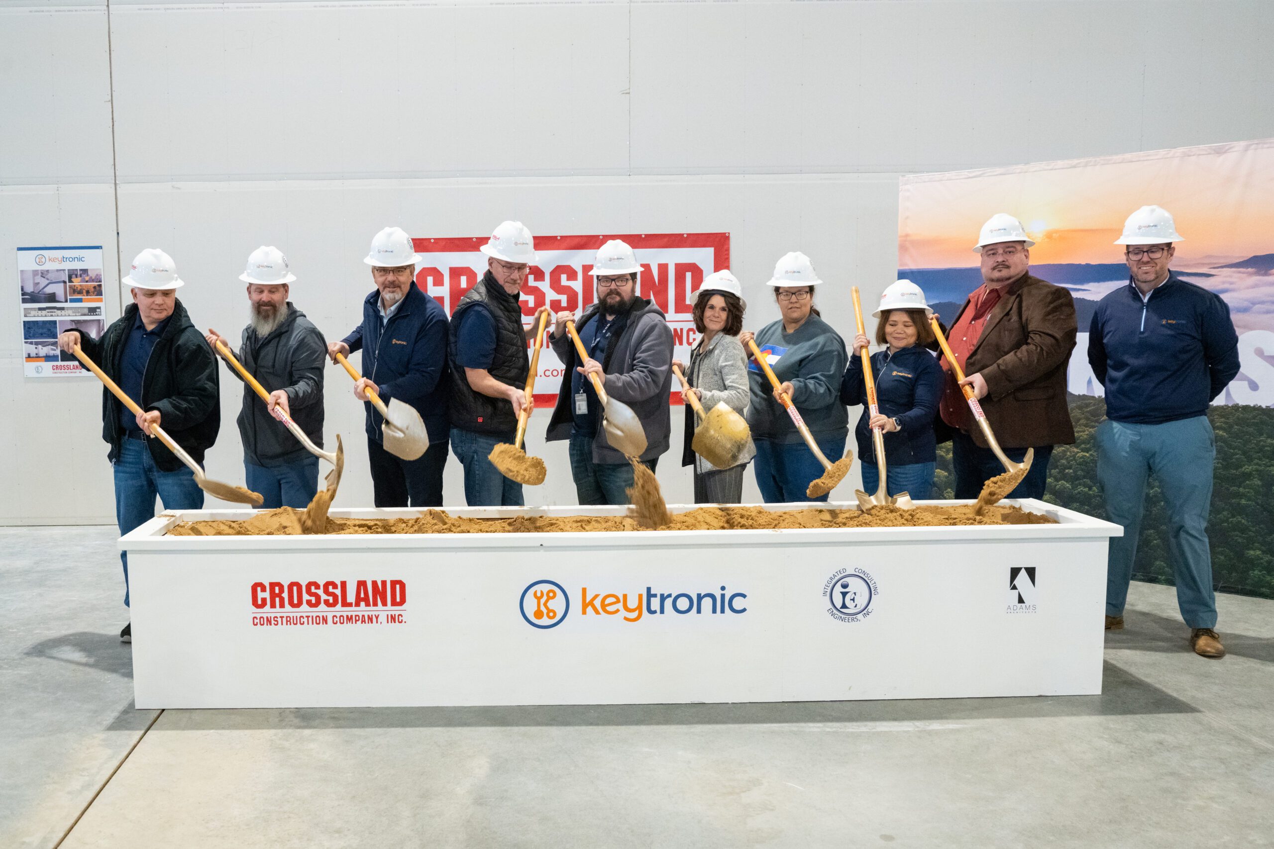 A group of ten people wearing hard hats and holding shovels participate in an indoor groundbreaking ceremony. They are standing behind a rectangular sand-filled box with the logos of Crossland Construction, Keytronic, Integrated Consulting Engineers, Inc., and Adams Architecture displayed on the front. A Crossland Construction banner is visible in the background, along with posters and a scenic backdrop. The participants are smiling and tossing sand with their shovels, celebrating the start of a new construction project.