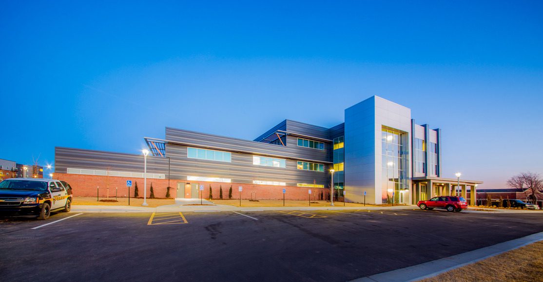 Modern three-story building with large windows and a mix of brick and metal siding, lit up at dusk. Several cars are parked in front, and the sky is clear and blue.