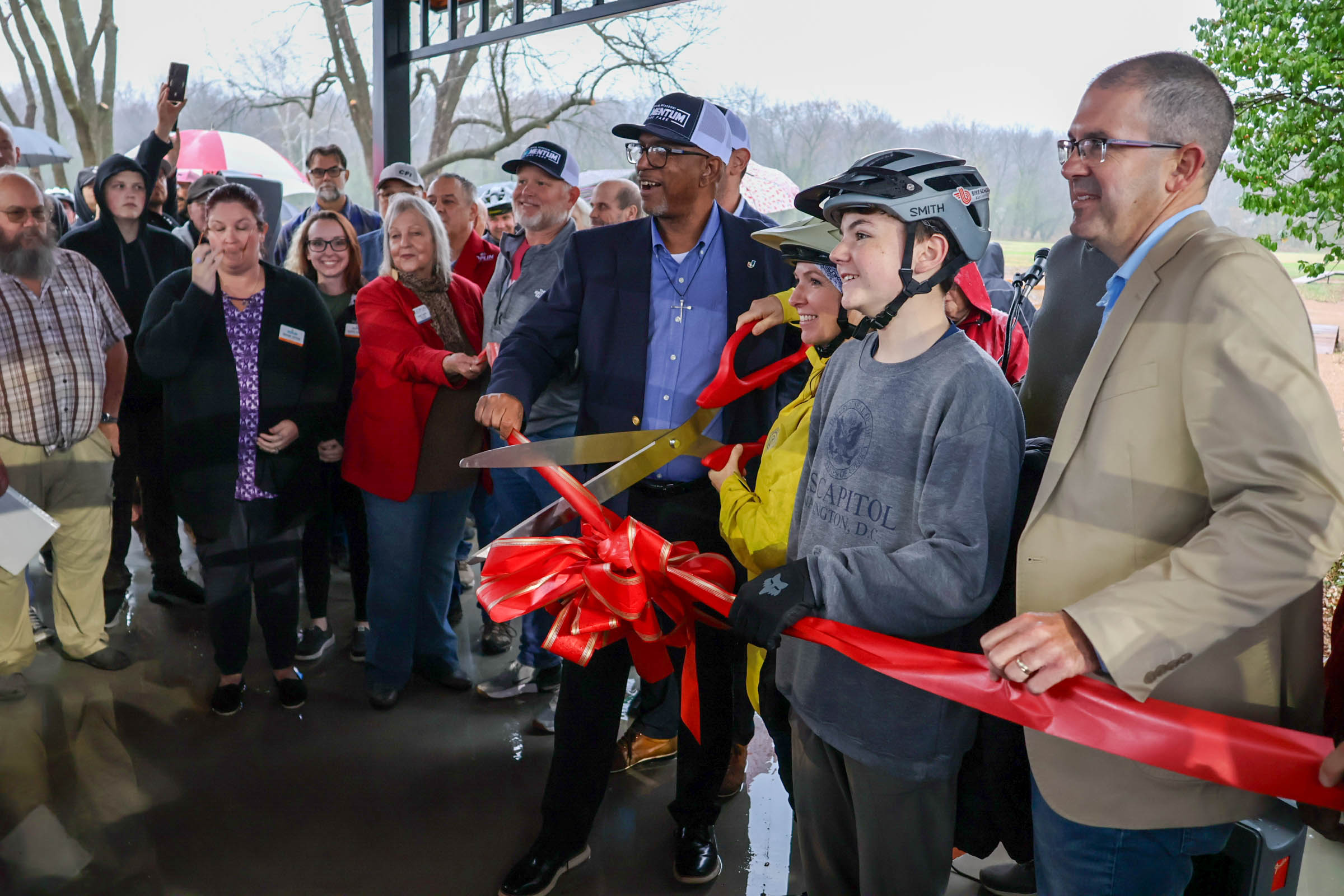 A group of people gather under a pavilion as a man in a suit and two young people wearing bike helmets cut a large red ribbon with oversized scissors, celebrating the opening of a new space or facility.