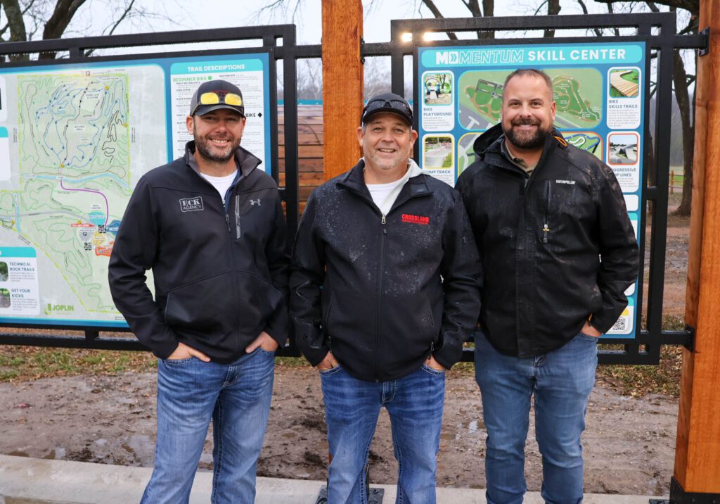 Three men wearing jackets and jeans stand smiling in front of large outdoor informational signs at the Momentum Skill Center. The ground is damp and trees without leaves are visible in the background.