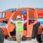 A Crossland Construction worker wearing a high-visibility vest and hard hat communicates with a colleague operating a Lull telehandler on a construction site. The operator, wearing a blue hard hat and red shirt, listens attentively from inside the cab. The bright orange telehandler, branded with the Crossland Construction logo, is positioned on a gravel surface with a scenic hill in the background.