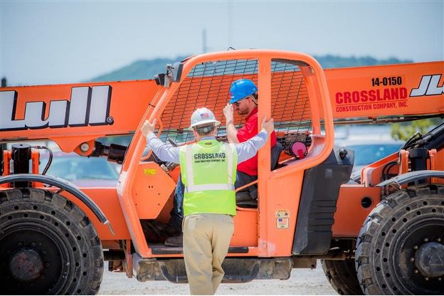 A Crossland Construction worker wearing a high-visibility vest and hard hat communicates with a colleague operating a Lull telehandler on a construction site. The operator, wearing a blue hard hat and red shirt, listens attentively from inside the cab. The bright orange telehandler, branded with the Crossland Construction logo, is positioned on a gravel surface with a scenic hill in the background.