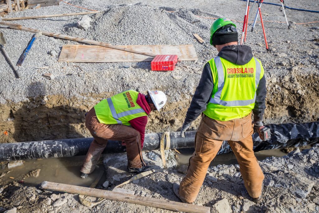Two construction workers in safety vests and helmets work on a large black pipe in a muddy trench at a construction site, surrounded by gravel and wooden planks.