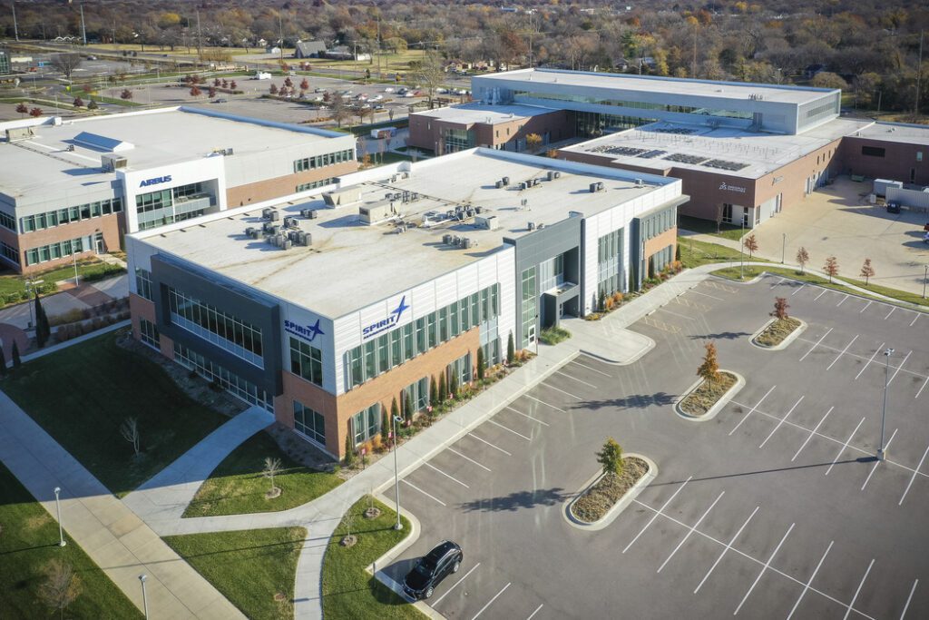 Aerial view of a modern corporate office complex with multiple brick and glass buildings, an empty parking lot, and landscaped grounds. Only one car is parked, and trees with autumn foliage surround the area.