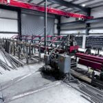 Metal fabrication workshop with industrial machinery, including a red RMS saw, metal bars, and partially assembled structures. The area is spacious with concrete floors and exposed metal beams.