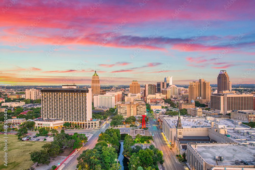 Aerial view of downtown San Antonio, Texas at sunrise, featuring tall buildings, green trees, and a colorful sky with pink and blue clouds.