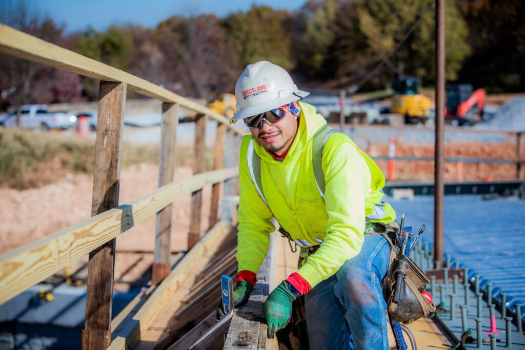 A construction worker in a neon yellow hoodie, hard hat, and safety gear kneels on a wooden structure at a building site, holding tools and looking at the camera. Trees and equipment are visible in the background.