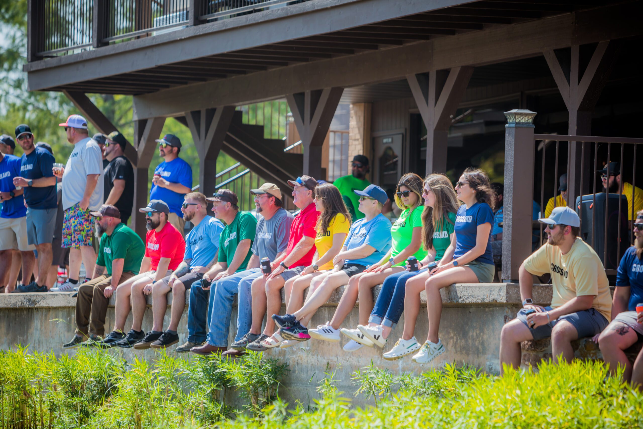 A group of people wearing colorful Crossland-branded t-shirts sit on a ledge outdoors, attentively watching an event. The participants appear engaged, with some wearing sunglasses and holding drinks. The background features a wooden deck with additional spectators standing and a staircase leading down to the gathering area. The setting is lush with greenery, indicating a relaxed, outdoor event.