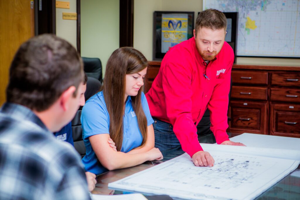 A man in a red shirt points to a large blueprint on a table while three colleagues listen and look on in a meeting room with maps and filing cabinets in the background.