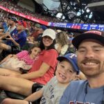 A smiling family of four wearing Texas Rangers gear sits in stadium seats at a baseball game, surrounded by other fans. The stands are full and the field is visible in the background.