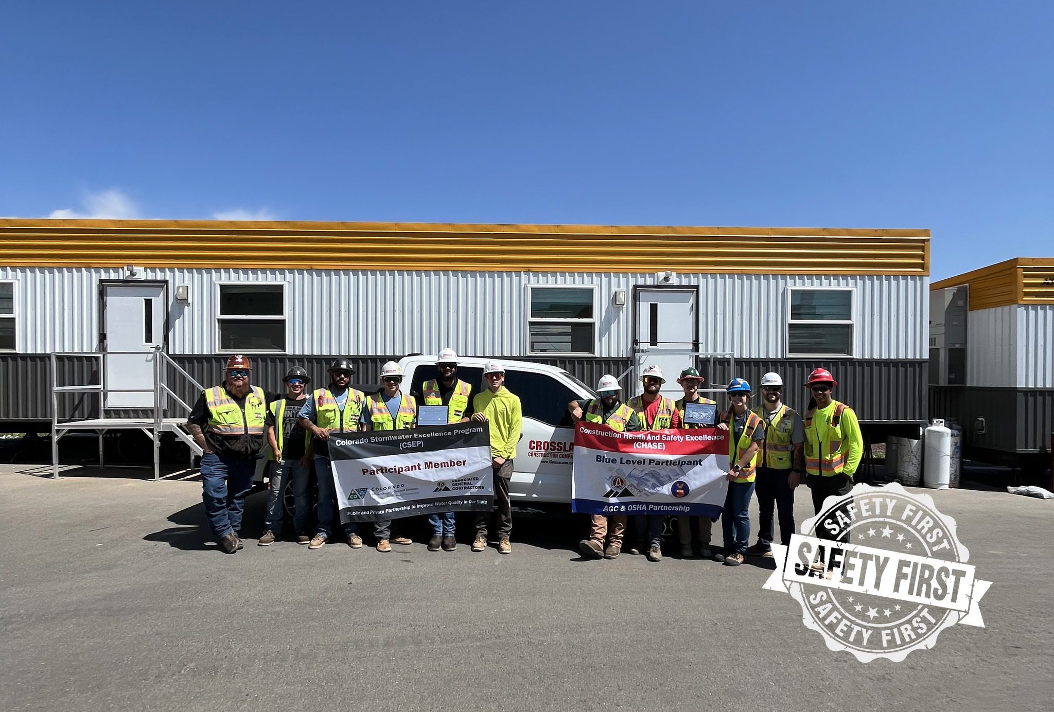 A group of construction workers wearing safety gear stand in front of trailers, holding banners promoting safety and partnership. A "Safety First" graphic is visible in the image’s bottom right corner.