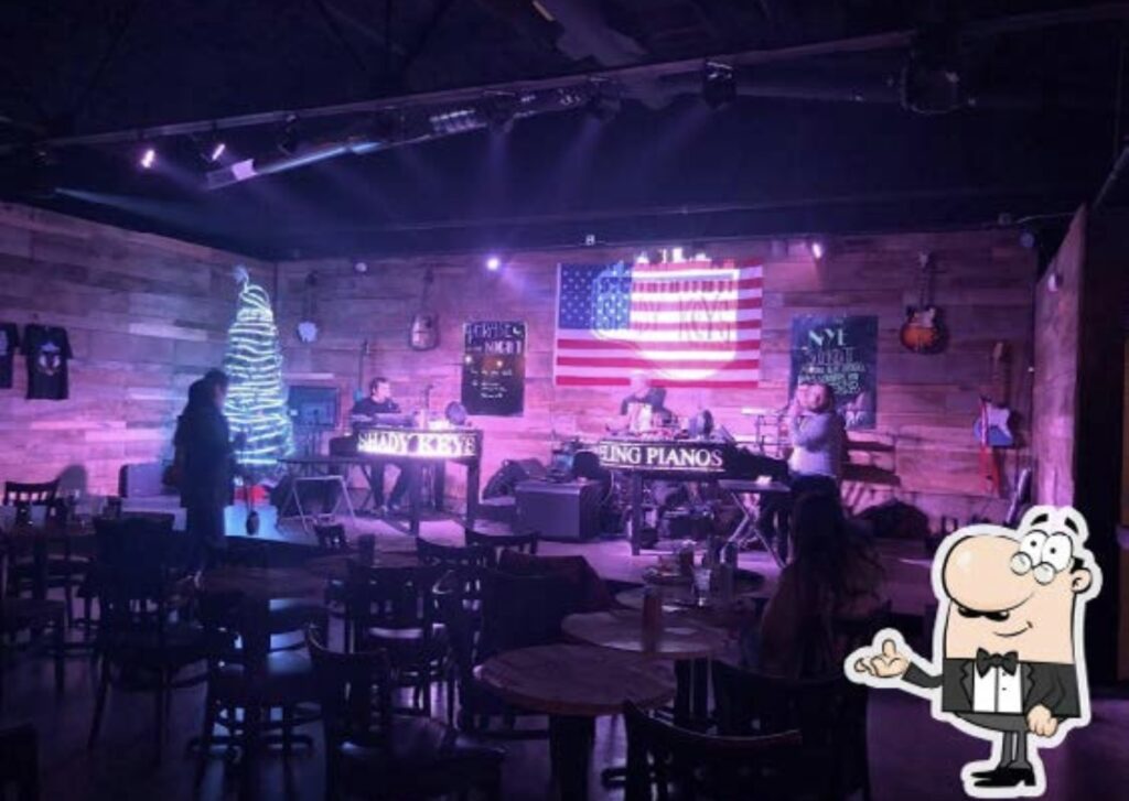 A band performs on stage in a dimly lit bar with wooden walls, an American flag, guitars on display, a lit-up Christmas tree, and tables and chairs scattered in the foreground.