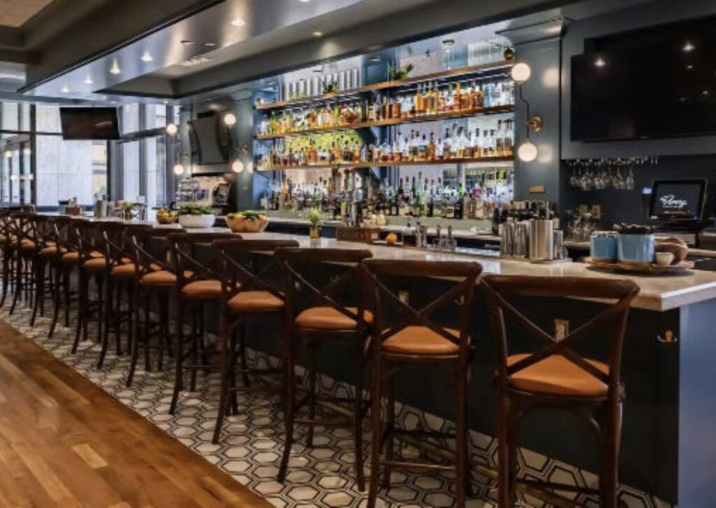 A modern bar with a long counter lined with wooden chairs, shelves stocked with bottles, hanging glasses, and mirrors behind the bar. The space has geometric floor tiles and two mounted TVs.