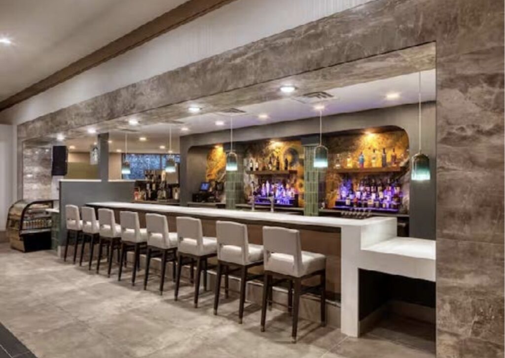 Modern bar with stone accents, a row of high-backed chairs, pendant lighting, and a well-stocked backlit liquor display. The setting is clean, stylish, and inviting with neutral tones and sleek design elements.