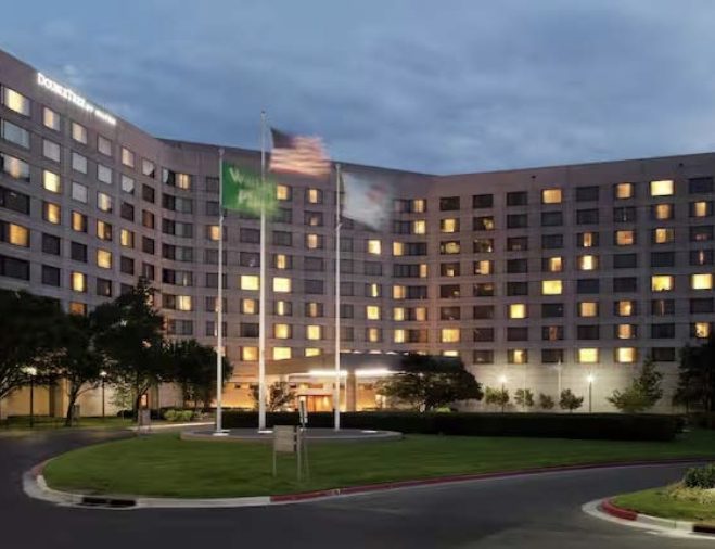 A large, multi-story hotel building with many lit windows at dusk, featuring three flagpoles in front and a circular driveway surrounded by greenery and trees.