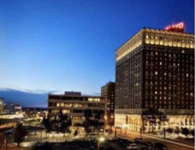 A cityscape at dusk featuring a tall, illuminated historic hotel with a red neon sign on its roof, surrounded by other buildings and streetlights. The sky is a deep blue, and cars are visible on the street below.