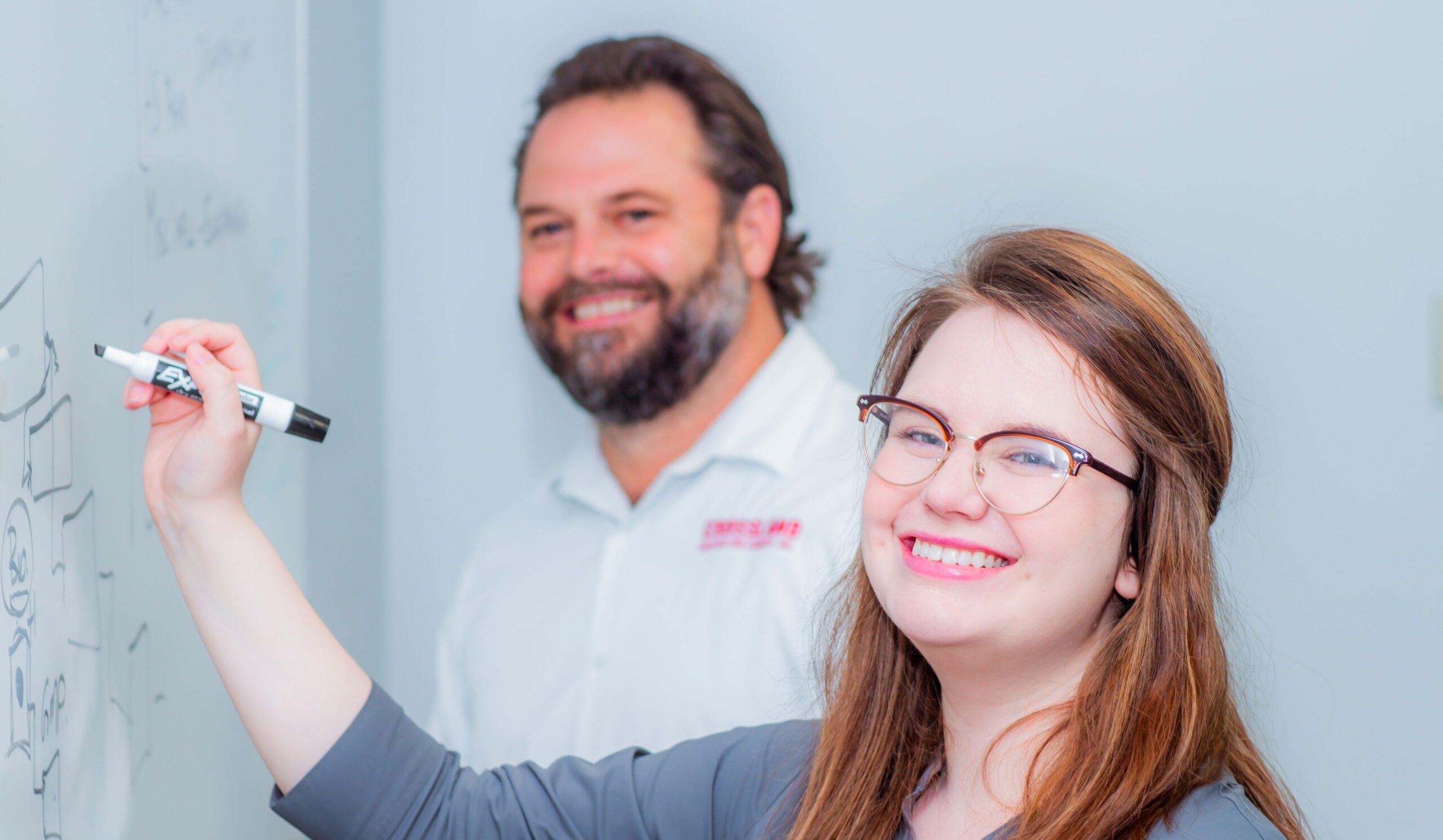 Two people writing on whiteboard and smiling at the camera.