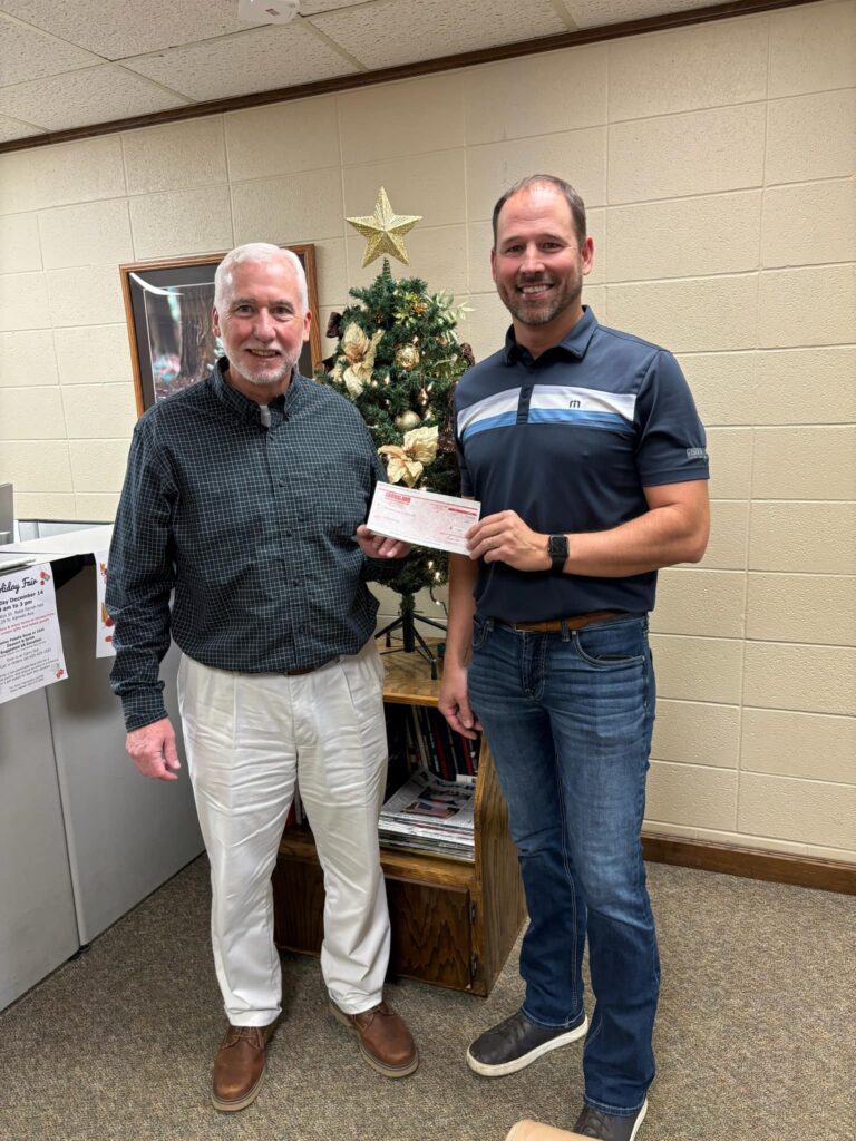 Two men stand indoors next to a small decorated Christmas tree. One is holding a check. Both are smiling at the camera. The wall behind them is beige with a framed photo and papers on a bulletin board.