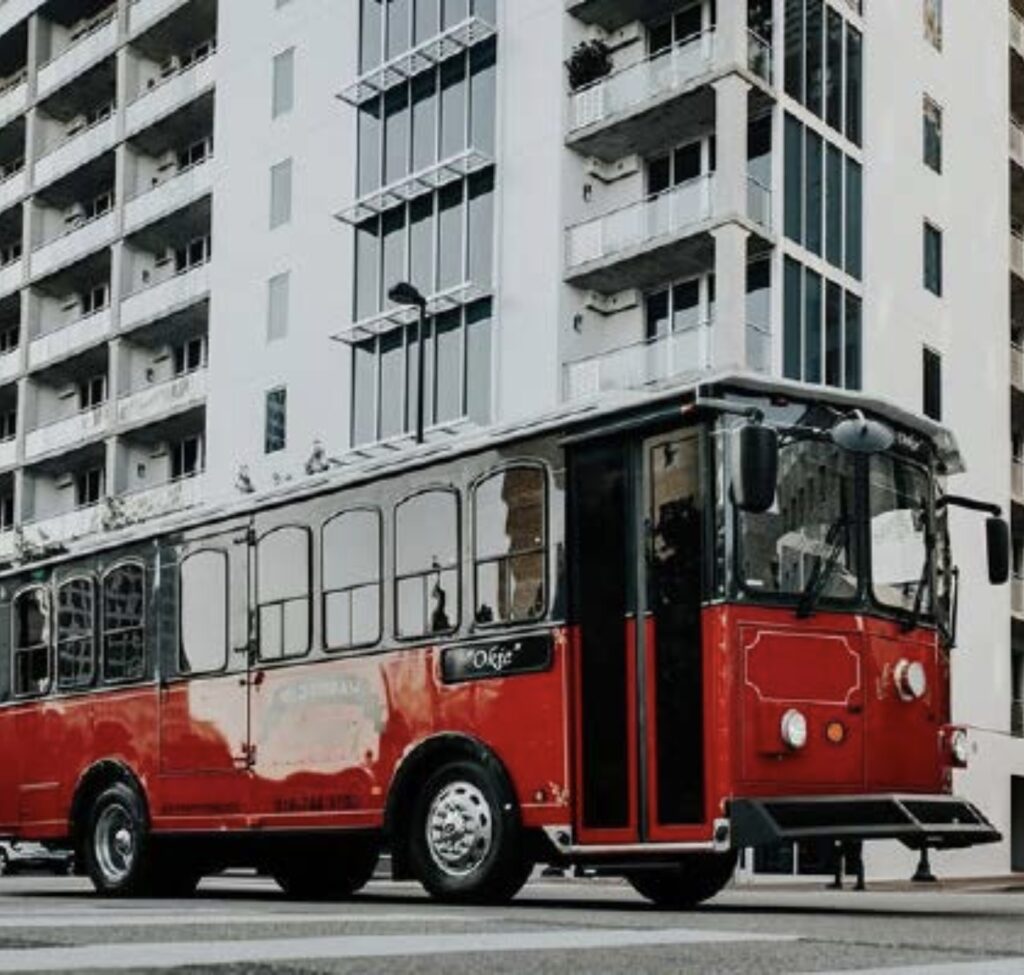 A red vintage-style trolley bus is parked on a city street in front of a modern multi-story apartment building.