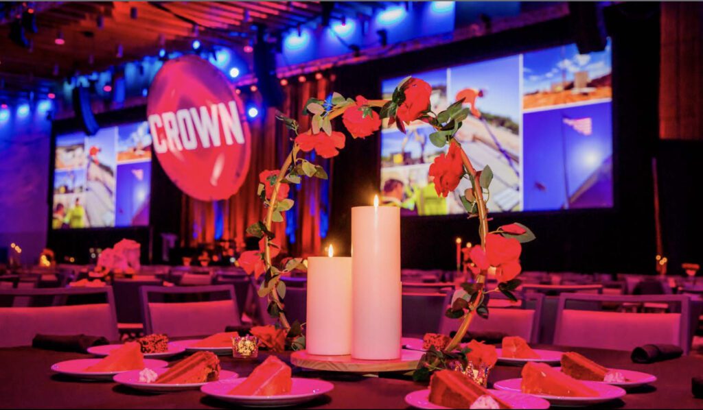 A centerpiece with three white candles and red roses sits on a table with plates of dessert, while a large screen displays images and the word "CROWN" at an event with vibrant lighting.
