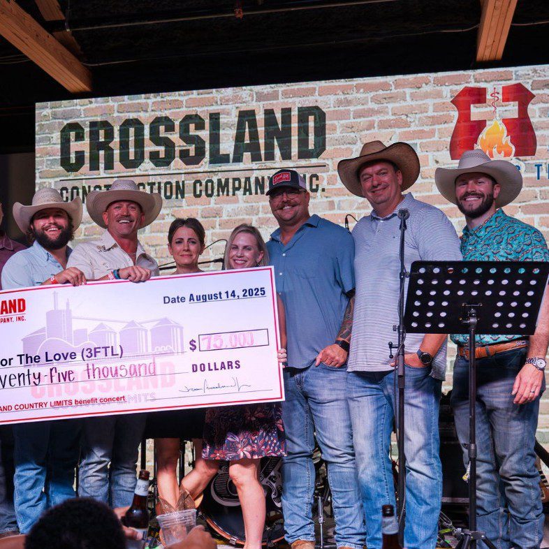 A group of seven people, some wearing cowboy hats, stand on a stage holding an oversized $25,000 check from Crossland Construction Company, dated August 14, 2025. A banner and microphone stand are visible in the background.