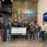 A group of people pose inside a food bank, holding a large check displaying "4,440 Pounds Packed" and a giant black fork. The food bank sign and a holiday tree are visible in the background.