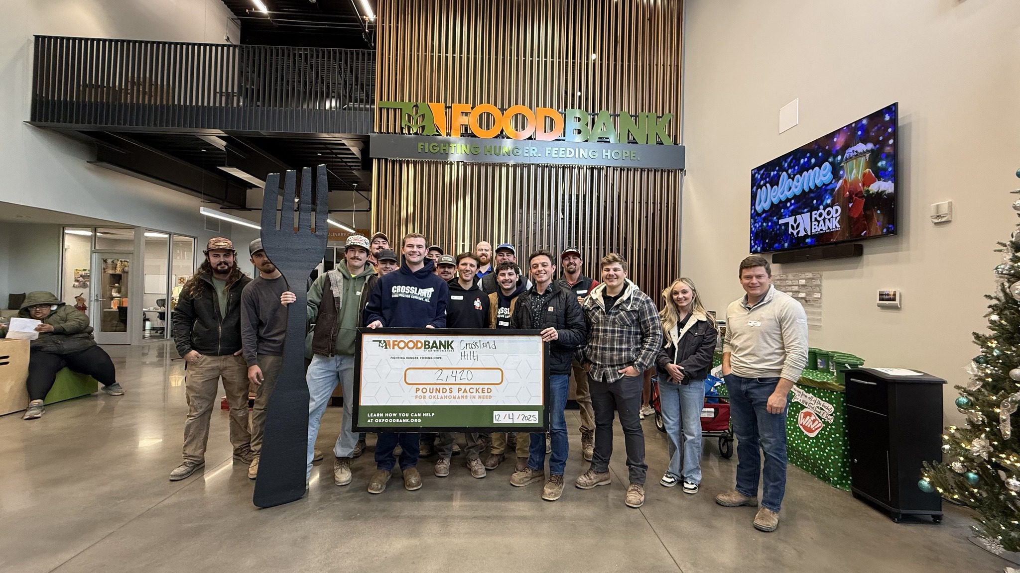 A group of people pose inside a food bank, holding a large check displaying "4,440 Pounds Packed" and a giant black fork. The food bank sign and a holiday tree are visible in the background.